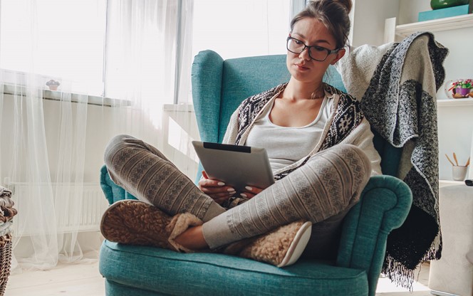 Woman By The Fireplace Sitting In A Cozy Armchair With A Warm Blanket Using A Tablet