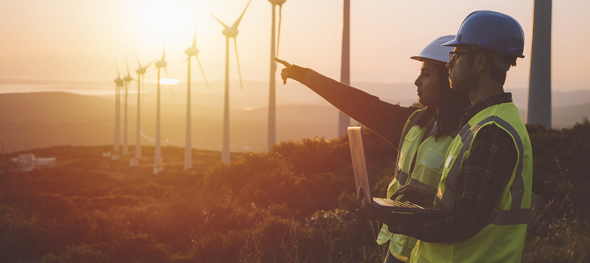Young Maintenance Engineer Team Working In Wind Turbine Farm At Sunset