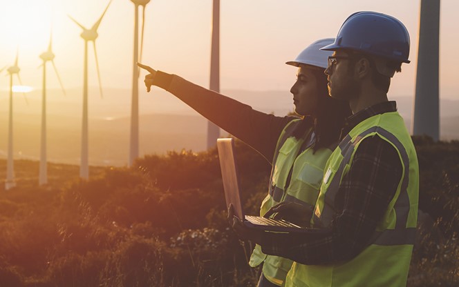 Young Maintenance Engineer Team Working In Wind Turbine Farm At Sunset