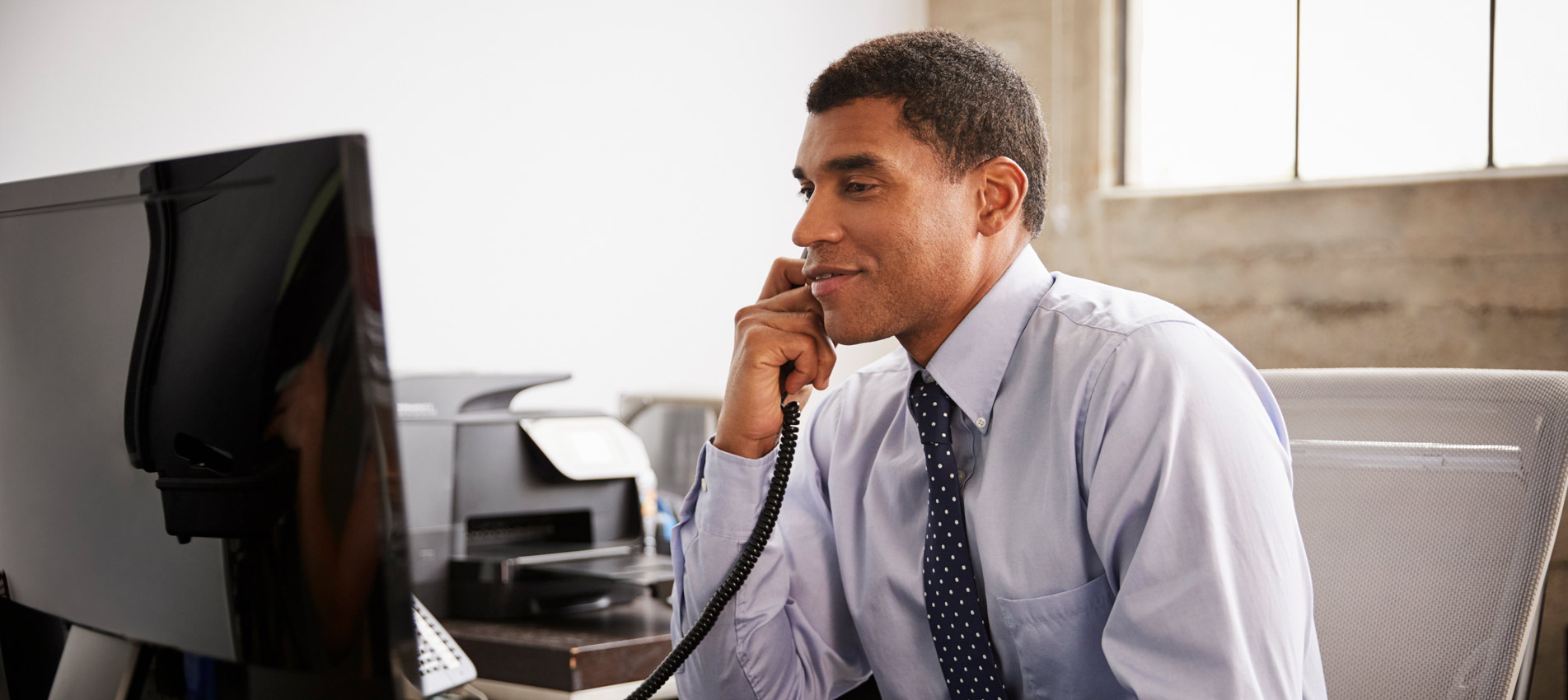 Businessman At An Office Desk Using Phone And Computer